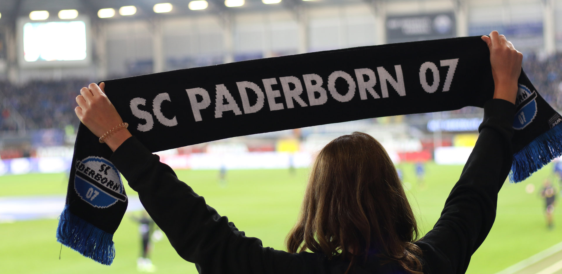 Im Vordergrund sieht man eine weibliche Person von hinten, die einen schwarzen Schal mit der weißen Aufschrift "SC Paderborn 07" in die Höhe hält. Im Hintergrund ist unscharf der Innenraum der Home Deluxe Arena zu erkennen.