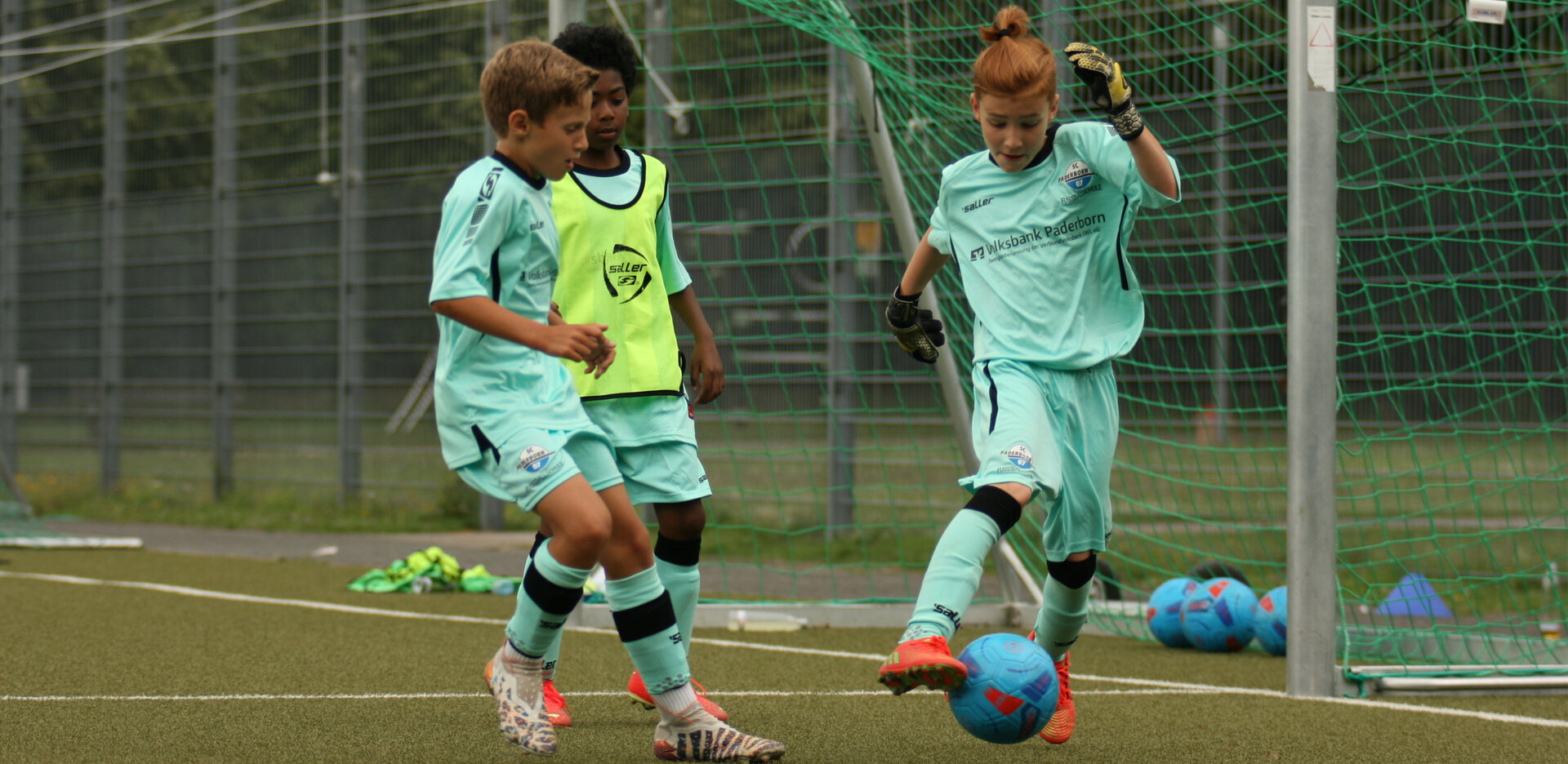 Drei Kinder in türkisen Trikotsets spielen Fußball und kämpfen um den Ball. Eines der Kinder trägt außerdem ein neongelbes Leibchen.