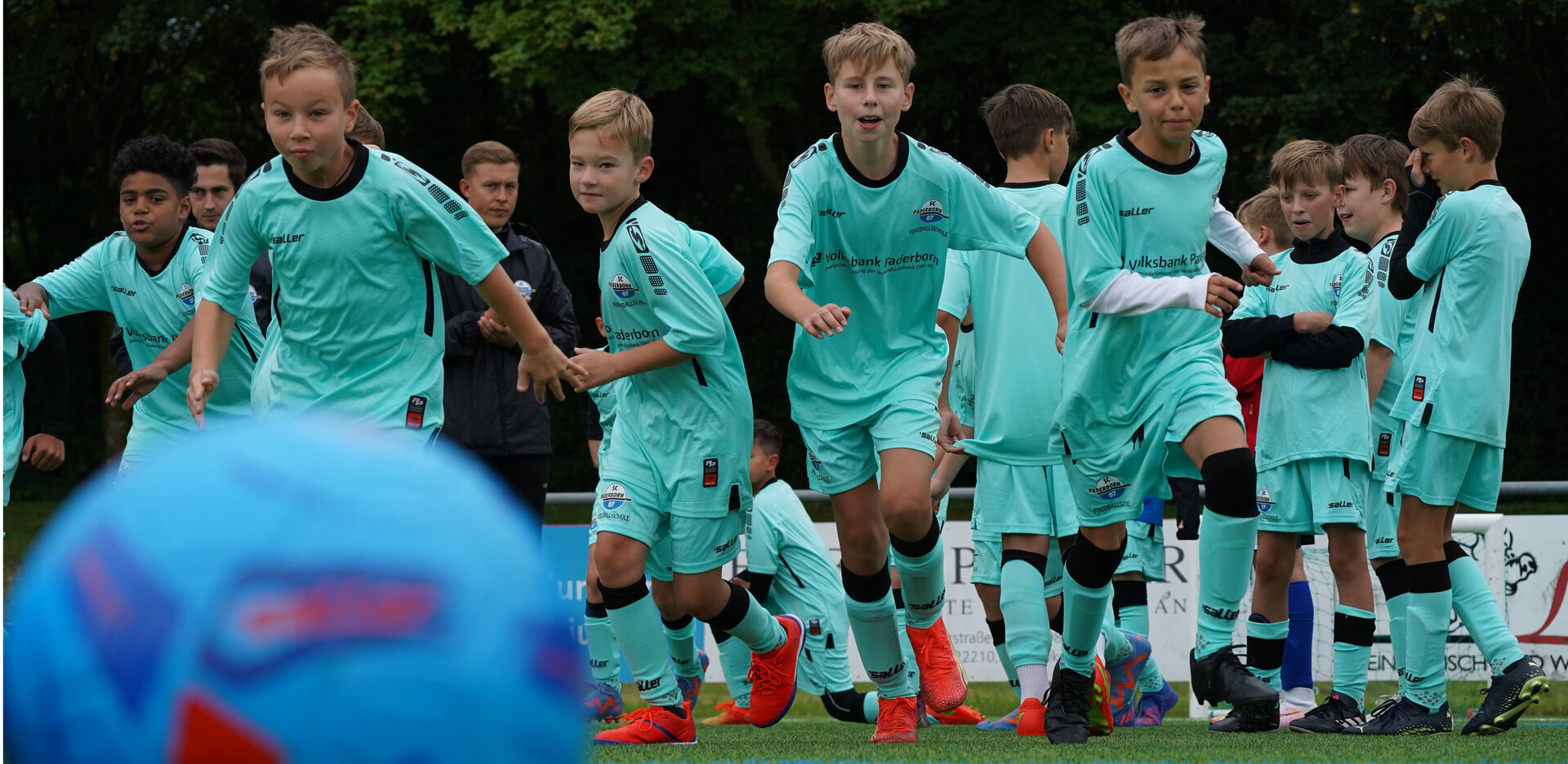 Einige Kinder in türkisen Trikotsets laufen auf einen im Vordergrund liegenden Ball zu.