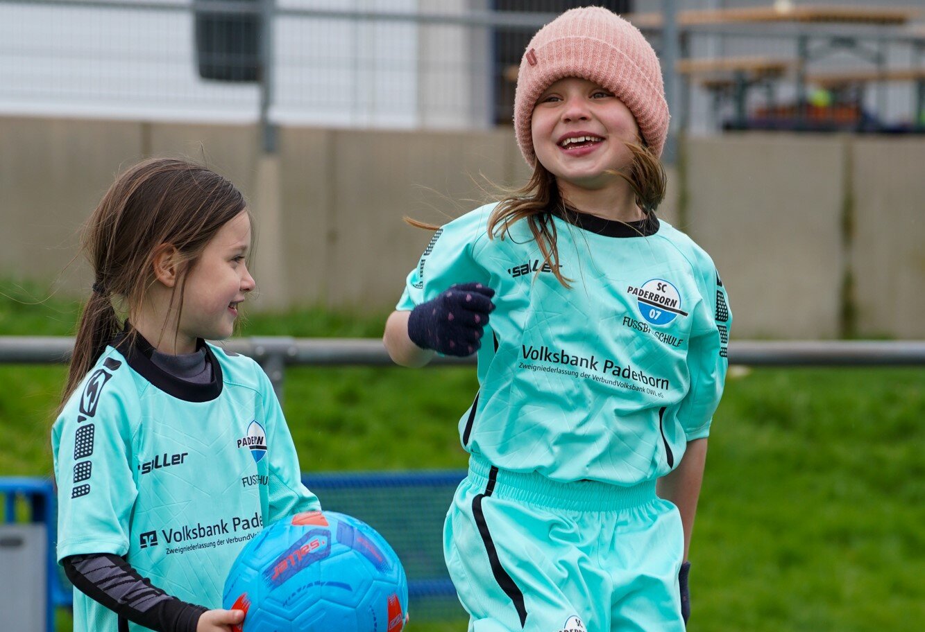 Zwei Mädchen mit türkisen Trikotsets beim Kickerkamp. Eines trägt eine rosa Mütze und lächelt in Richtung Kamera, das andere hat einen Ball in der Hand und schaut das Mädchen mit der Mütze an.