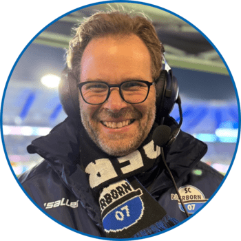 Porträt von Reporter Daniel Prior in der Home Deluxe Arena. Er trägt kurze blonden Haare, eine runde Brille mit dunklem Rahmen, einen Fünf-Tage-Bart sowie einen SCP07-Schal über seiner dunkelblauen Winterjacke.