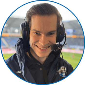 Porträt von Reporter Felix Willmes in der Home Deluxe Arena. Er trägt kurze dunkelblonde Haare, keinen Bart sowie eine geöffnete dunkelblaue Winterjacke unter der ein dunkelblauer Pullover sichtbar ist.