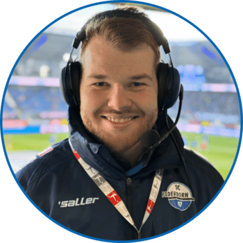 Porträt von Reporter Henry Lausen in der Home Deluxe Arena. Er hat kurze rötliche Haare, einen Sieben-Tage-Bart und trägt ein Bundesliga-Schlüsselband über seiner dunkelblauen Winterjacke.