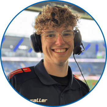 Porträt von Reporter Julian Thies in der Home Deluxe Arena. Er trägt seine blonden, mittellangen Haare lockig, eine runde Brille sowie ein dunkelblaues Polohemd.