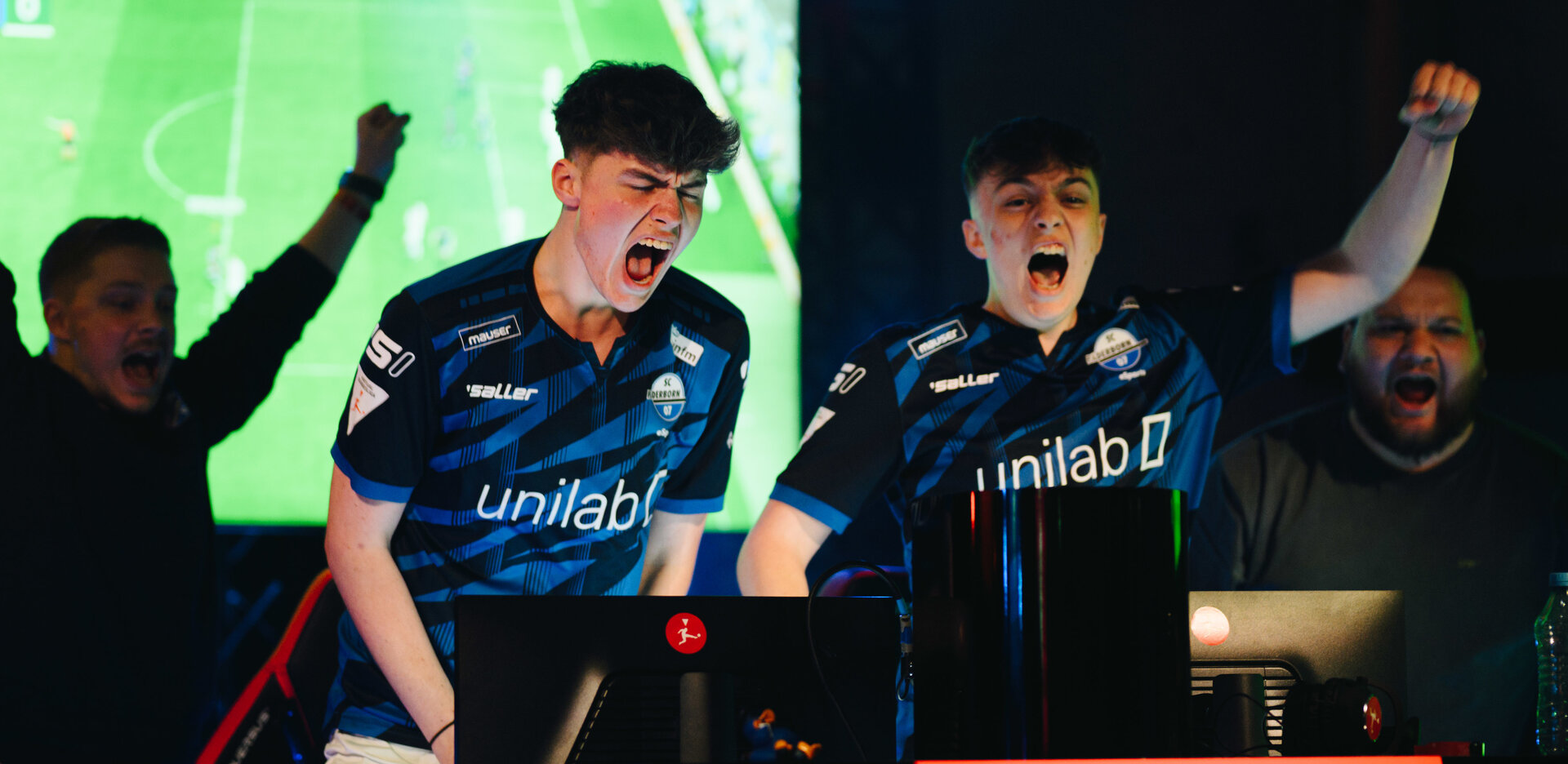 Im Vordergrund jubeln die beiden eSportler "Jonny" und "Chaser" beim Gewinn der deutschen Club-Meisterschaft hinter ihren Bildschirmen. Dahinter jubelt auch Trainer "Coach_Pommes", er ist jedoch aufgrund eines Bildschirms hinter ihm nur als Silhouette erkennbar.