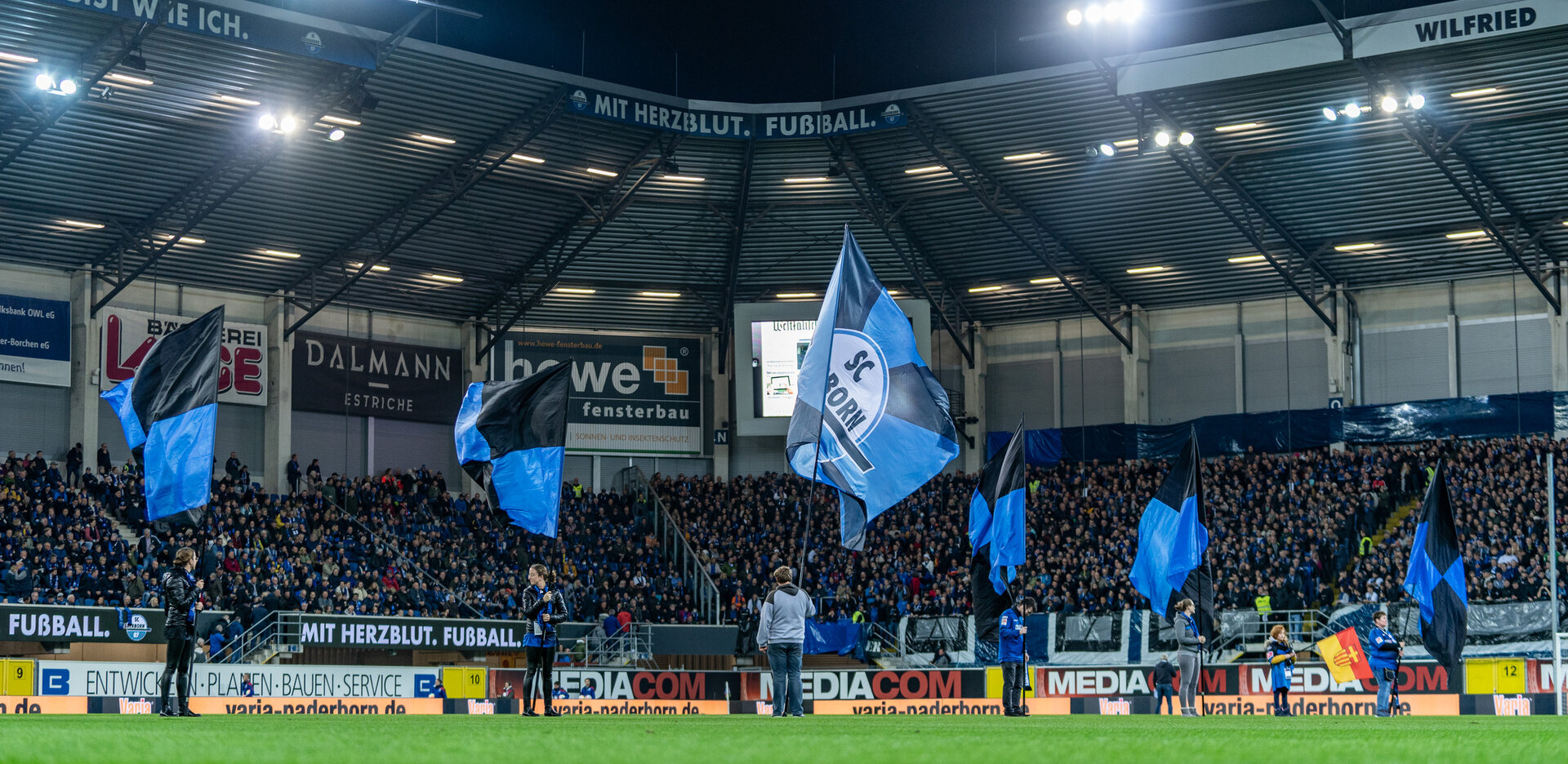 Bei einem Flutlichtspiel in der Home Deluxe Arena stehen fünf Fahnenschwenker auf dem Rasen und ihre blau-schwarzen SCP07-Fahnen wehen im Wind.